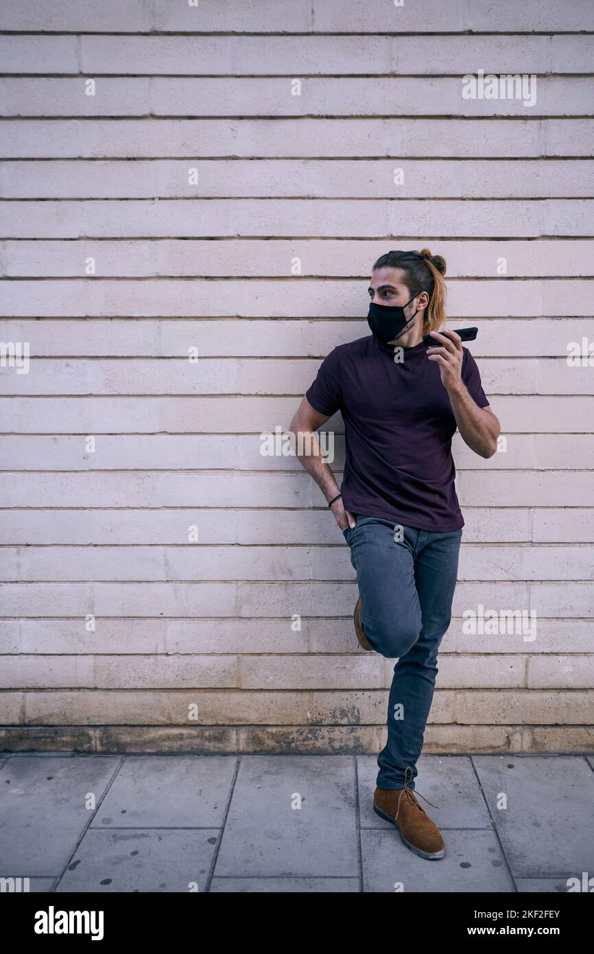 caucasian guy with long beard hair standing in black t-shirt leaning on dirty city wall with smartphone in left hand wearing face mask Stock Photo