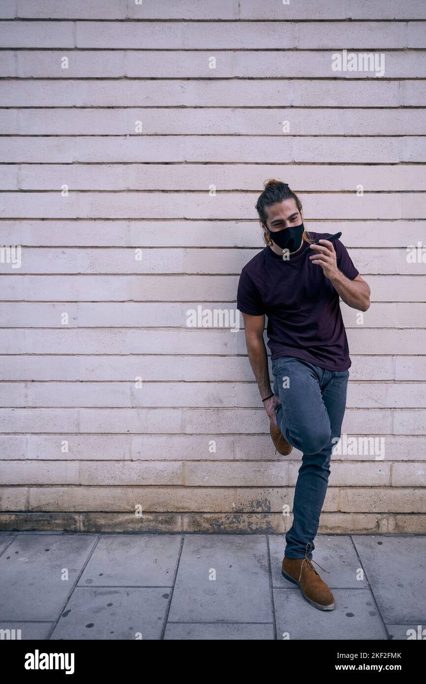 caucasian guy with long beard hair standing in black t-shirt leaning on the dirty wall in the city talking on the smartphone with face mask Stock Photo