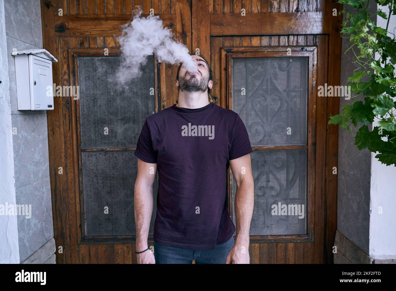 caucasian young man with beard exhaling water vapor through his mouth near wooden door and mailbox Stock Photo