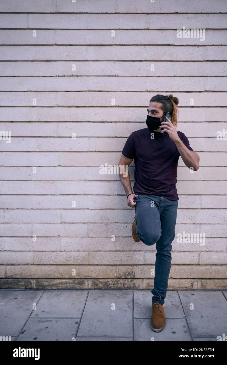 caucasian guy standing in black t-shirt brown shoes leaning on the dirty wall in the city talking on the smartphone wearing face mask Stock Photo