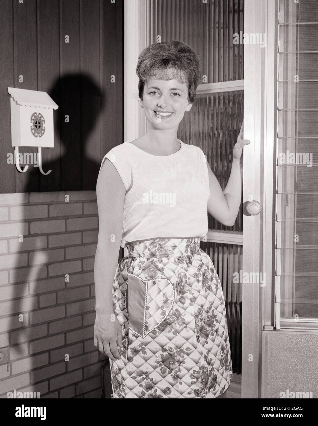 1960s SMILING HOUSEWIFE WEARING APRON STANDING WITH FRONT DOOR AJAR LOOKING AT CAMERA - s14156 HAR001 HARS FRIENDSHIP HALF-LENGTH LADIES PERSONS RESIDENTIAL BUILDINGS ANSWERING B&W EYE CONTACT HOMEMAKER HAPPINESS HOMEMAKERS CHEERFUL DISCOVERY PRIDE HOMES HOUSEWIVES SMILES WELCOMING AJAR JOYFUL RESIDENCE MID-ADULT MID-ADULT WOMAN BLACK AND WHITE CAUCASIAN ETHNICITY HAR001 OLD FASHIONED Stock Photo