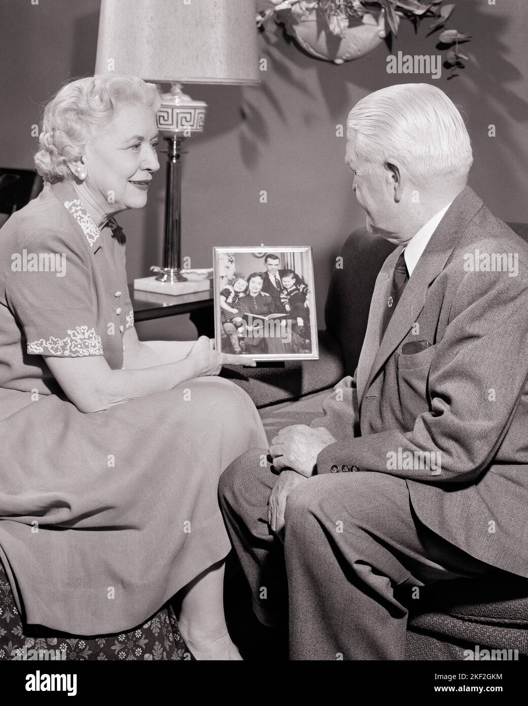 1950s SENIOR COUPLE SITTING IN LIVING ROOM LOOKING AT FRAMED PHOTO OF THEIR CHILDREN AND GRANDCHILDREN - s357 HAR001 HARS GRANDPARENTS FAMILIES JOY LIFESTYLE ELDER FEMALES MARRIED GRANDPARENT STUDIO SHOT SPOUSE HUSBANDS HOME LIFE COPY SPACE FRIENDSHIP HALF-LENGTH LADIES PERSONS INSPIRATION MALES RETIREMENT SENIOR MAN GRANDCHILDREN SENIOR ADULT B&W PARTNER SENIOR WOMAN RETIREE HAPPINESS OLD AGE OLDSTERS OLDSTER AND PRIDE TO GRANDMOTHERS K ELDERS CONNECTION GRANDDAUGHTER GRANDFATHERS FRAMED GRANDSON ELDERLY MAN ELDERLY WOMAN TOGETHERNESS WIVES BLACK AND WHITE CAUCASIAN ETHNICITY GRANDMA GRANDPA Stock Photo