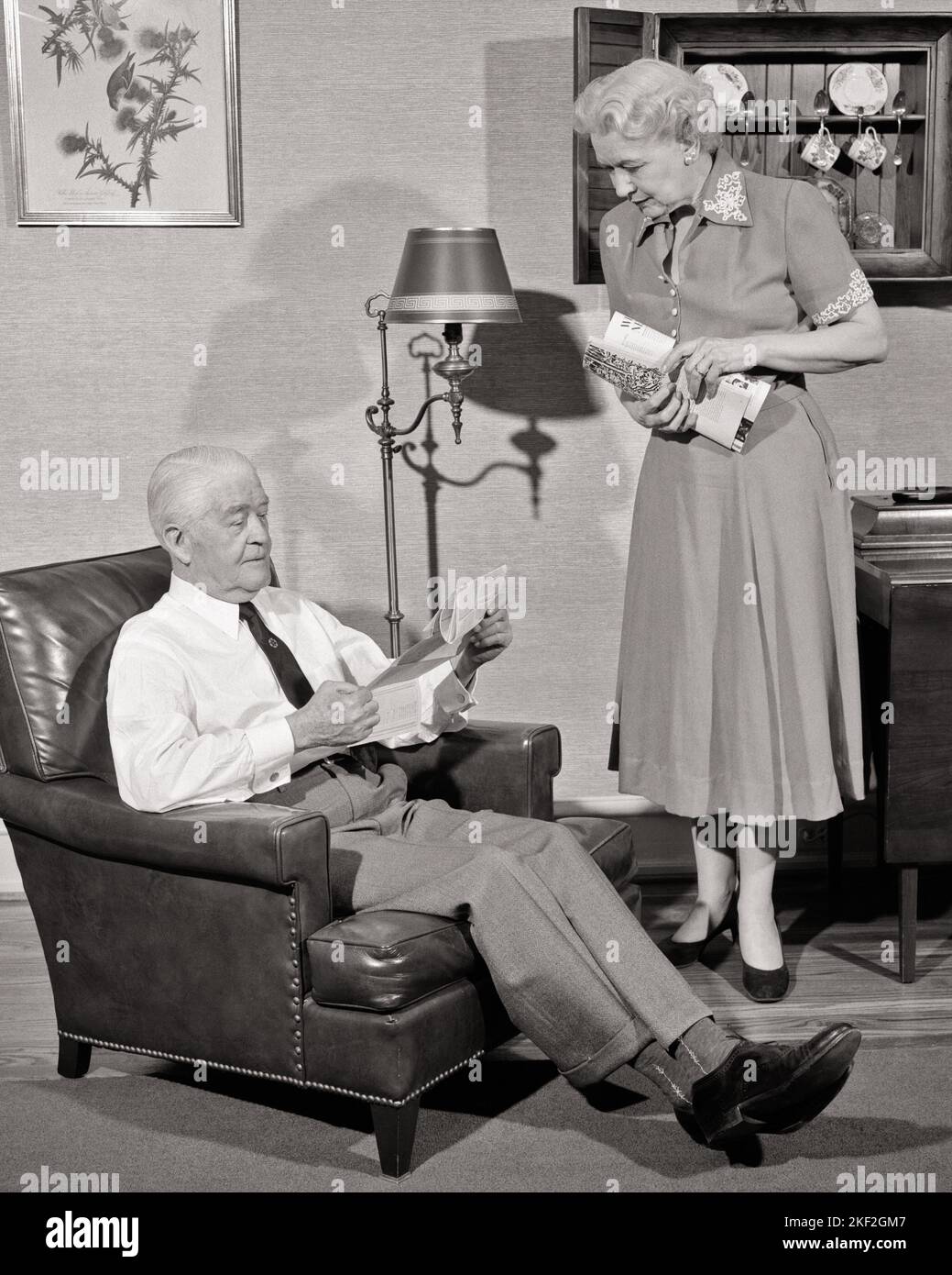 1950s SENIOR MAN AND WOMAN AT HOME WIFE STANDING CARRYING BUNDLE OF MAIL HUSBAND SITTING IN CHAIR READING LETTER  - s390 HAR001 HARS OLD FASHION RETIRED 1 COMMUNICATION BALANCE TEAMWORK LIFESTYLE ELDER FEMALES MARRIED SPOUSE HUSBANDS HOME LIFE COPY SPACE FULL-LENGTH LADIES PERSONS CARING MALES RETIREMENT SENIOR MAN SENIOR ADULT B&W PARTNER SENIOR WOMAN RETIREE OLD AGE OLDSTERS OLDSTER LIVING ROOM AND AT IN OF ELDERS CONCEPTUAL STYLISH BUNDLE ELDERLY MAN GRAY HAIR RELAXATION TOGETHERNESS WIVES AT HOME BLACK AND WHITE CAUCASIAN ETHNICITY HAR001 OLD FASHIONED Stock Photo