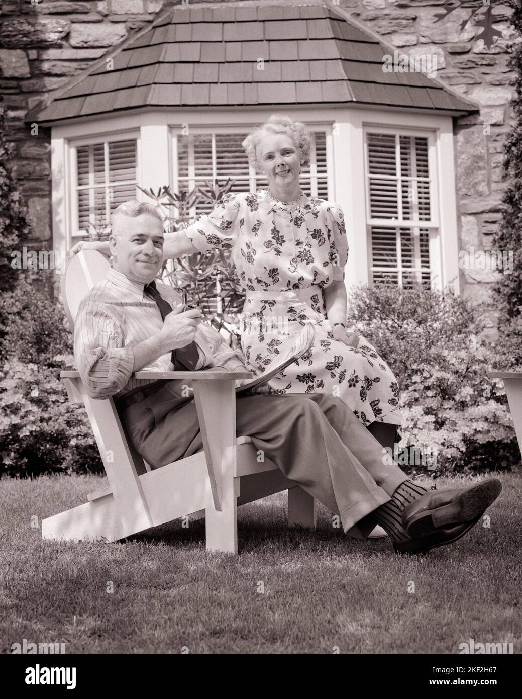 1940s PORTRAIT SENIOR COUPLE SITTING ADIRONDACK STYLE CHAIR ON FRONT LAWN STONE HOME WITH BAY WINDOW SMILING LOOKING AT CAMERA - s9435 HAR001 HARS DESIGN PLEASED JOY LIFESTYLE SATISFACTION ARCHITECTURE ELDER HOUSES PIPE BAY HEALTHINESS HOME LIFE LUXURY COPY SPACE FRIENDSHIP HALF-LENGTH RESIDENTIAL BUILDINGS RETIREMENT CONFIDENCE SENIOR MAN SENIOR ADULT ADIRONDACK MIDDLE-AGED B&W MIDDLE-AGED MAN SENIOR WOMAN SUCCESS RETIREE STRUCTURE HAPPINESS OLD AGE MIDDLE-AGED WOMAN OLDSTERS CHEERFUL OLDSTER LEISURE EXTERIOR PRIDE HOMES SMILES ELDERS JOYFUL RESIDENCE STYLISH COOPERATION TOGETHERNESS Stock Photo