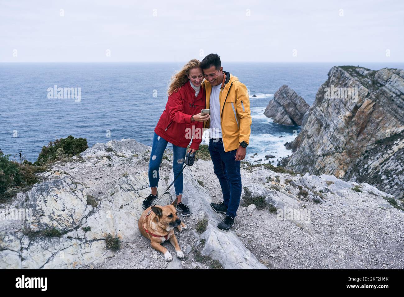 blonde caucasian girl standing on the rocks near the sea next to a dog showing her smartphone screen to a latin guy with yellow jacket, cabo de penas Stock Photo