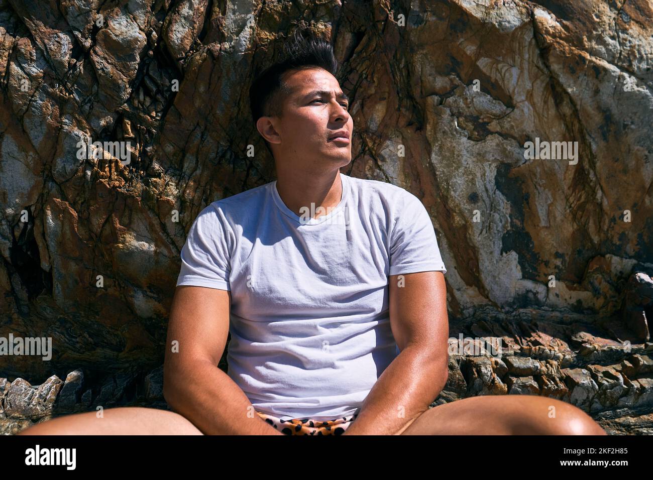 bust of a dark-haired latin boy with a swimsuit and blue t-shirt sitting on a large rock calm and relaxed looking to the side on a sunny day, asturias Stock Photo