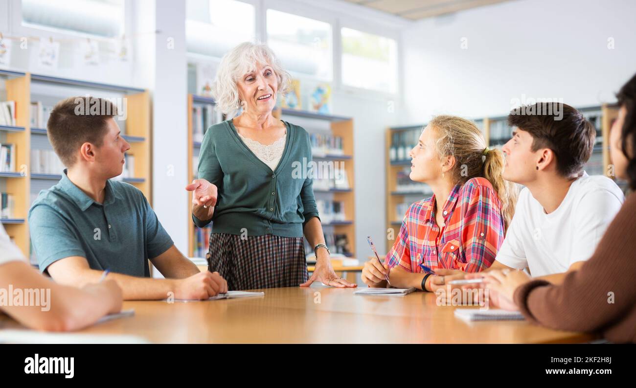 Elderly teacher, together with teenagers students, conducts table classes in school library Stock Photo