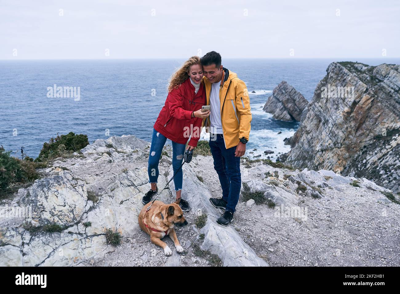 blonde caucasian girl with big dog lying on the ground and latin guy having fun on the rocks near the sea looking at the smartphone screen, cabo de Stock Photo