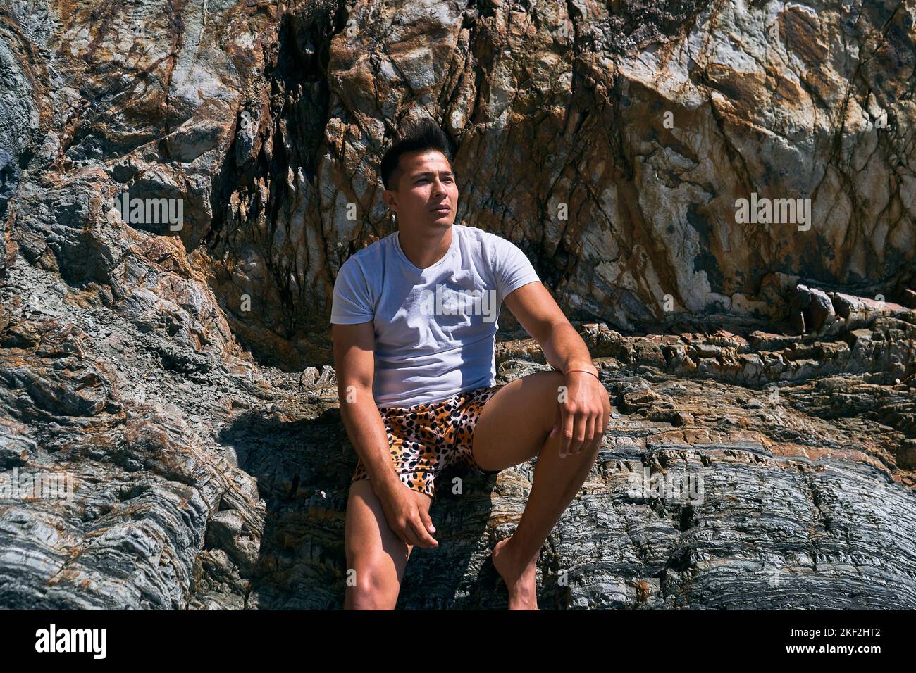 dark-haired latin boy with swimming trunks and blue t-shirt barefoot sitting on a big rock calm and relaxed on a sunny day, asturias, spain Stock Photo
