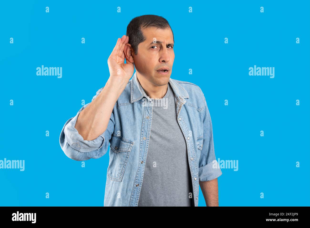Studio shot of a handsome  man with over isolated blue background smiling with hand over ear listening and hearing to rumor or gossip. Deafness concep Stock Photo