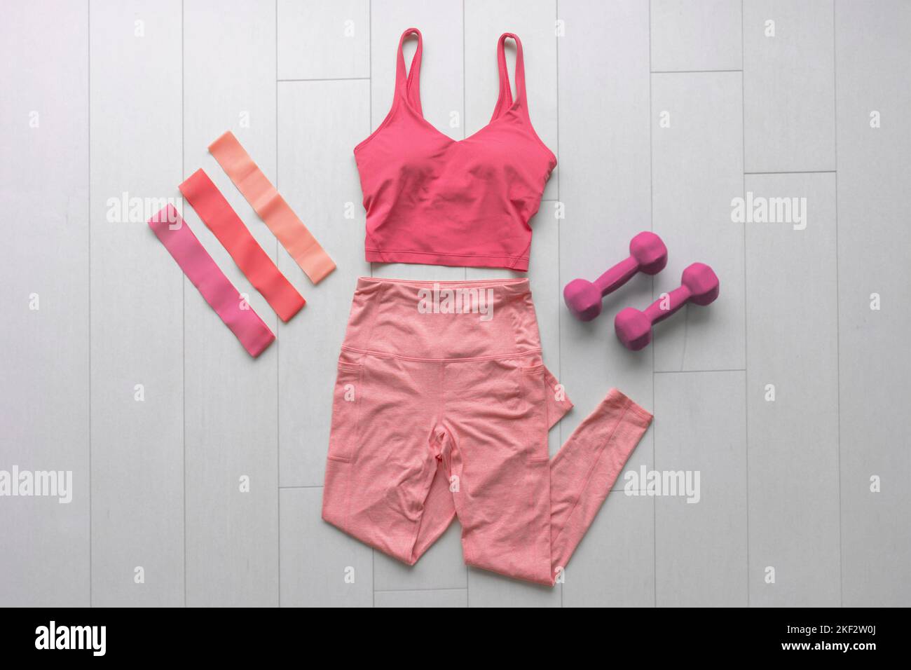 Fitness clothes flat lay workout at home with resistance bands and dumbbell weights. Pink athleisure fashion clothing top view on white wood floor Stock Photo
