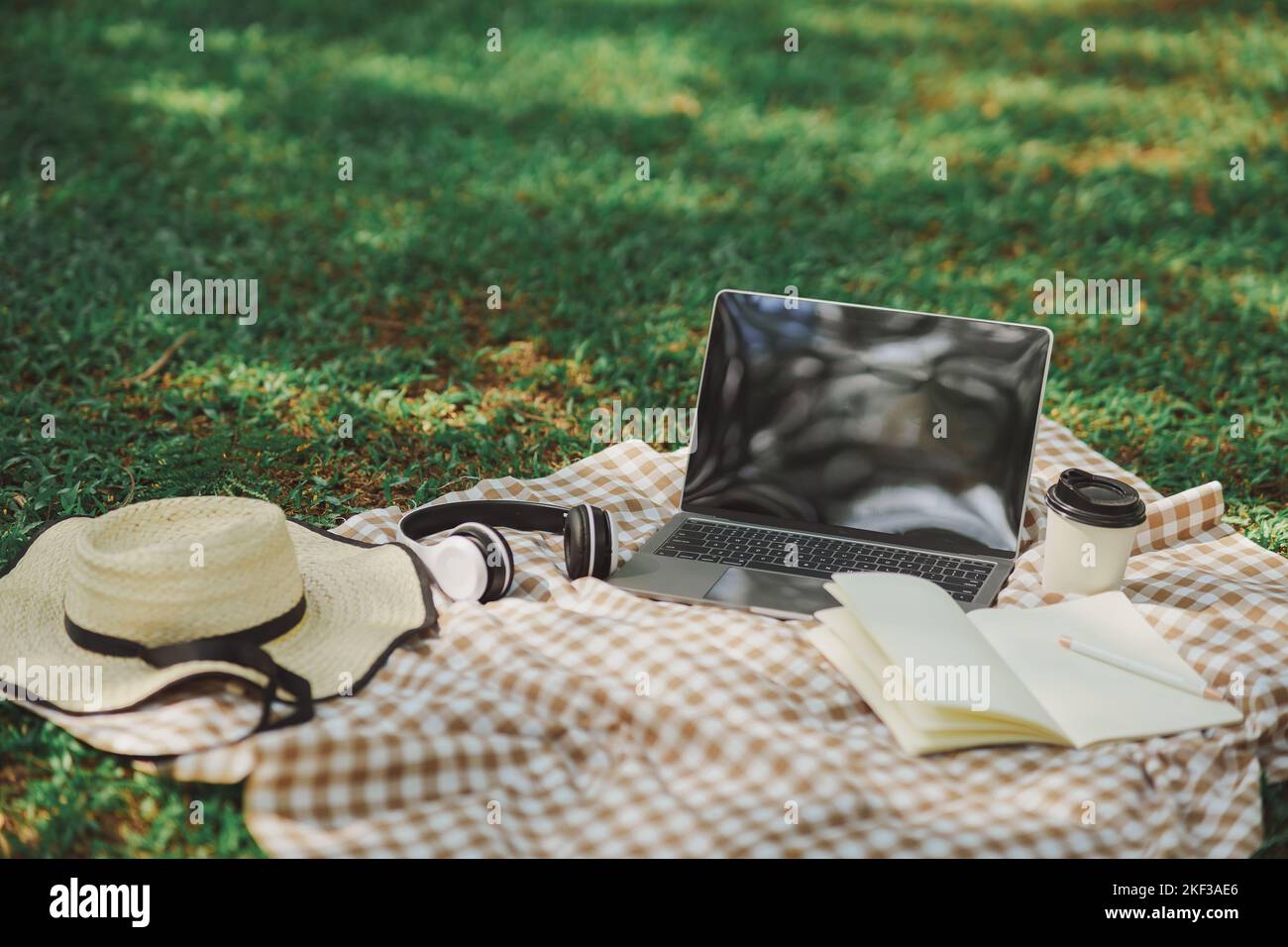 Work on laptop on picnic blanket on the lawn in the park- next to a headphone, notebook, pen, hat and cup of hot coffee. Freelancer work concept. Top Stock Photo