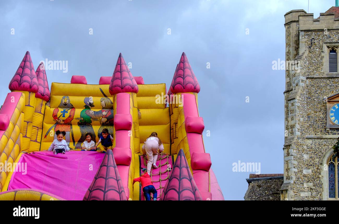 Children play on large inflatable medieval castle slide next to 14th Century Grade II* listed Church at Pinner Fair, June, 2022. Stock Photo