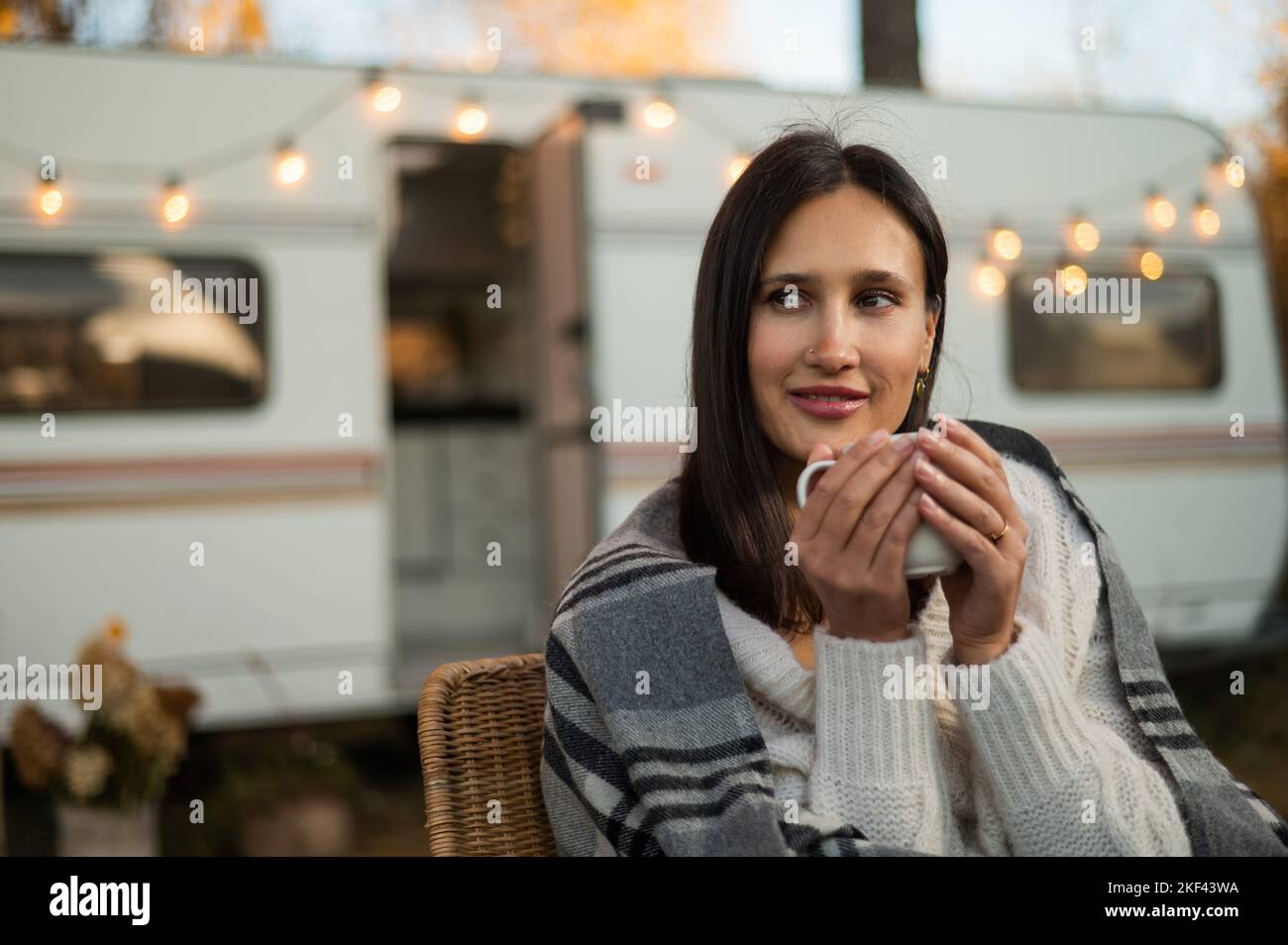 Caucasian woman in a knitted hat wrapped in a plaid and drinks a warming drink outdoors. Travel in a motor home in the fall. Stock Photo
