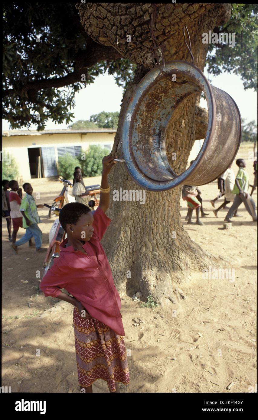Burkina Faso, Fada region; Girl ringing the schoolbell that hangs from a branch. Stock Photo