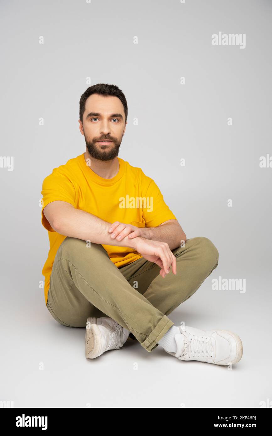 full length of man in yellow t-shirt and beige pants sitting with crossed legs on grey background,stock image Stock Photo