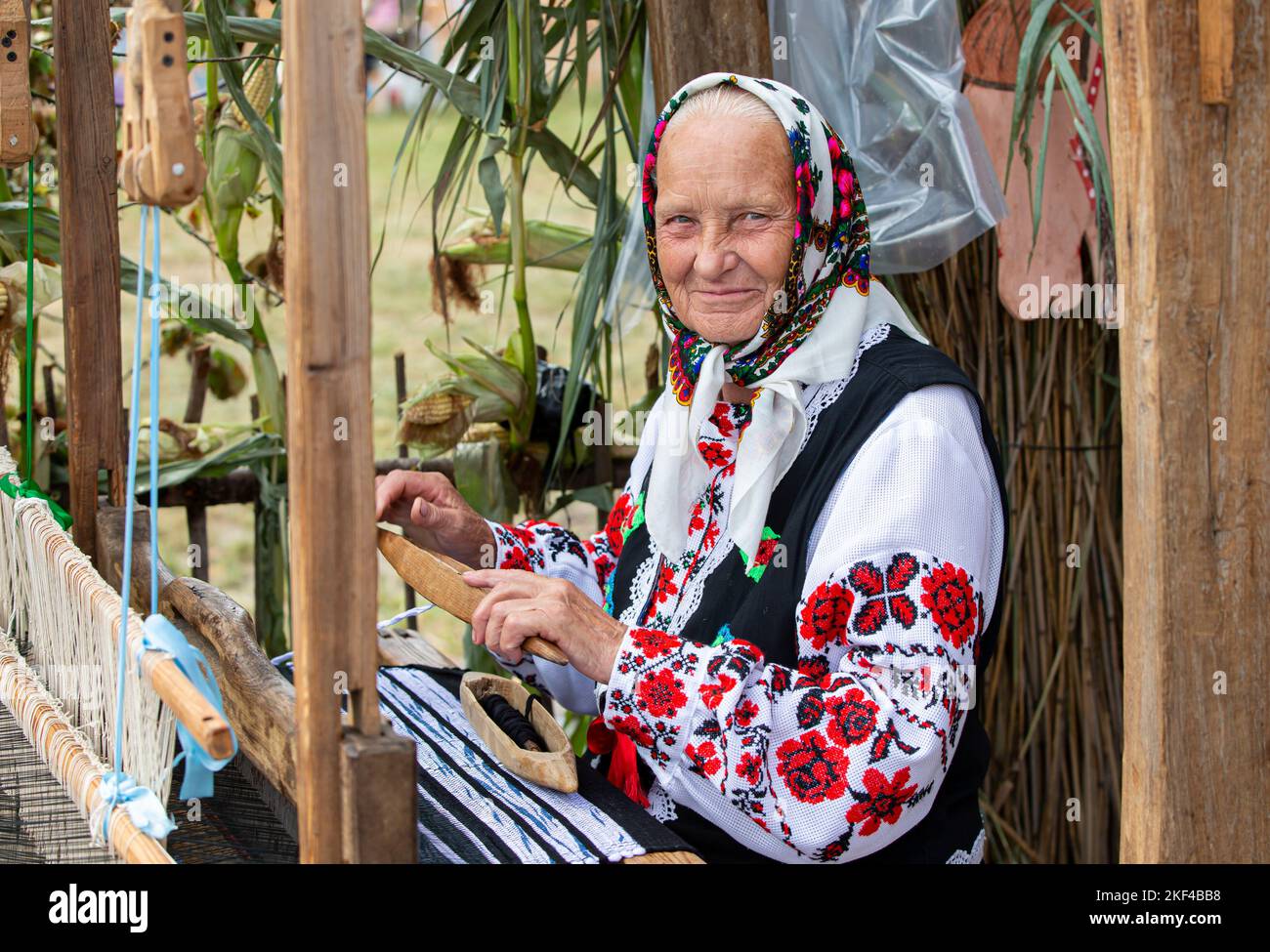 An old Belarusian or Ukrainian woman in an embroidered shirt at a vintage loom. Slavic elderly woman in national ethnic clothes. Stock Photo