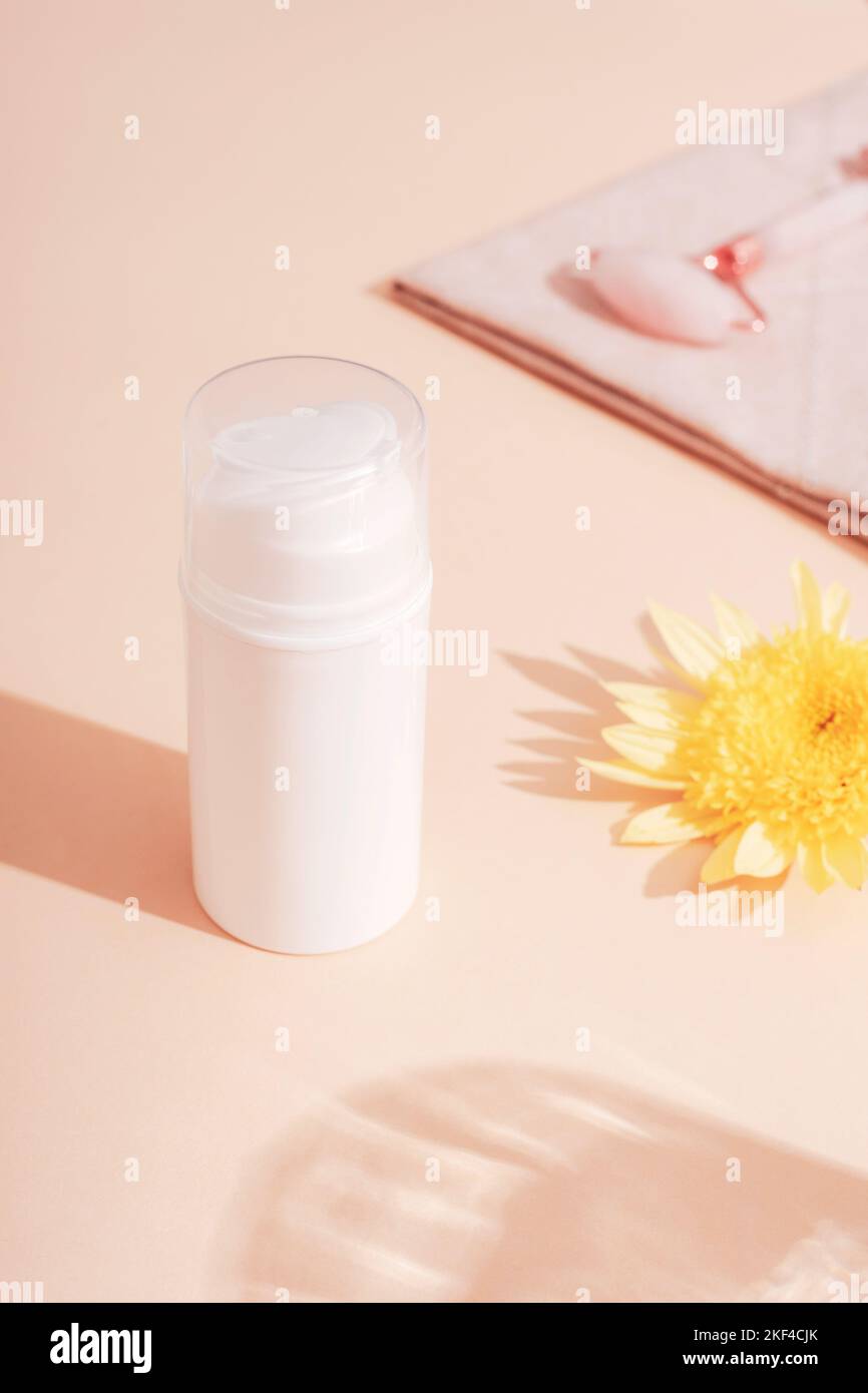 White cosmetic bottle, yellow flower and facial roller on beige background in sunlight. Skincare, spa and wellness concept. Stock Photo