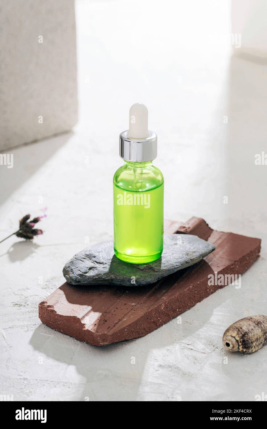 Green cosmetic serum bottle on natural stones podium in sunlight. Front view, mockup. Stock Photo