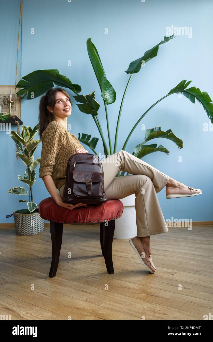 beautiful slender girl in beige pants and olive sweater posing while sitting with a small brown leather backpack Stock Photo