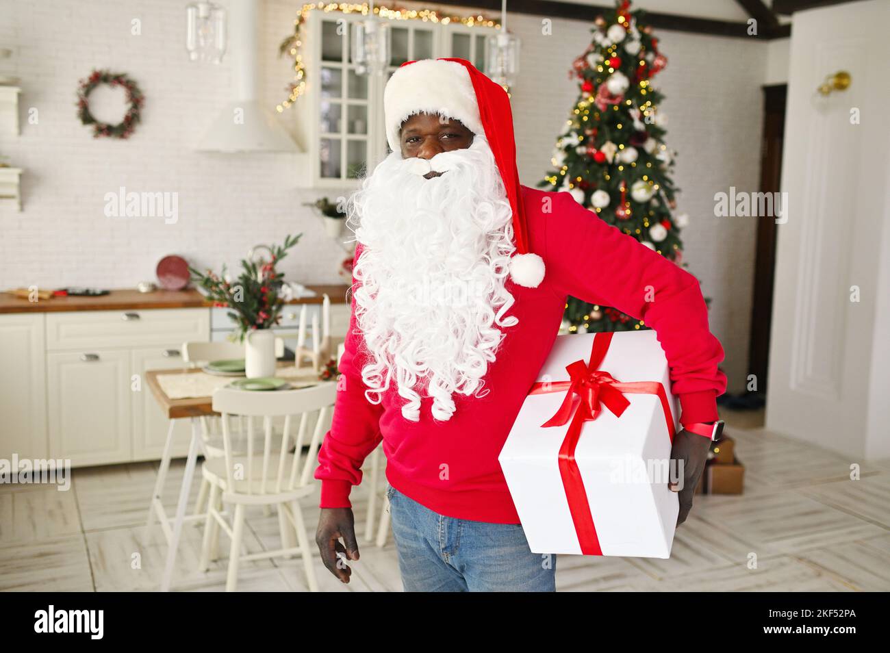african american man in a red sweater with a white false beard and a santa hat smiles and holds a large white box with a red ribbon in his hands on Stock Photo