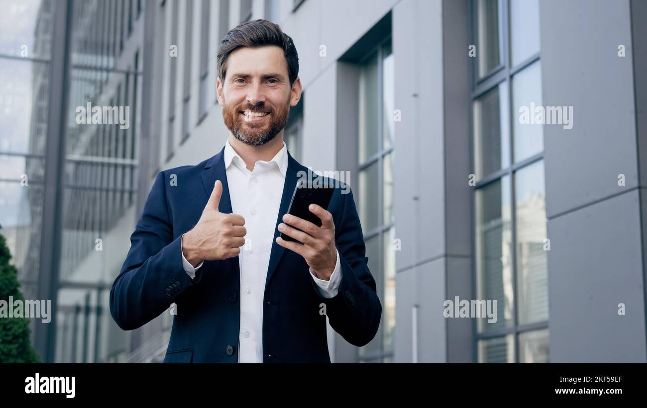 Happy man standing outdoors holding phone smiling businessman using new mobile app for business tasks showing thumb up gesture of approval Stock Photo