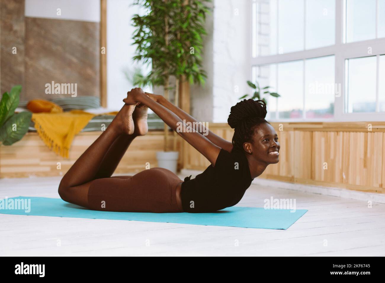 Beautiful and athletic afro american woman lying with smile in bow pose side view, doing yoga on gymnastic mat in decorated studio. Muscle stretching Stock Photo