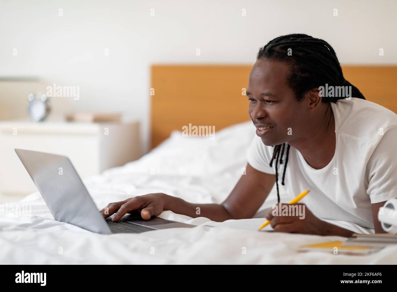 Online Education Concept. Black Young Man Using Laptop While Lying In Bed Stock Photo