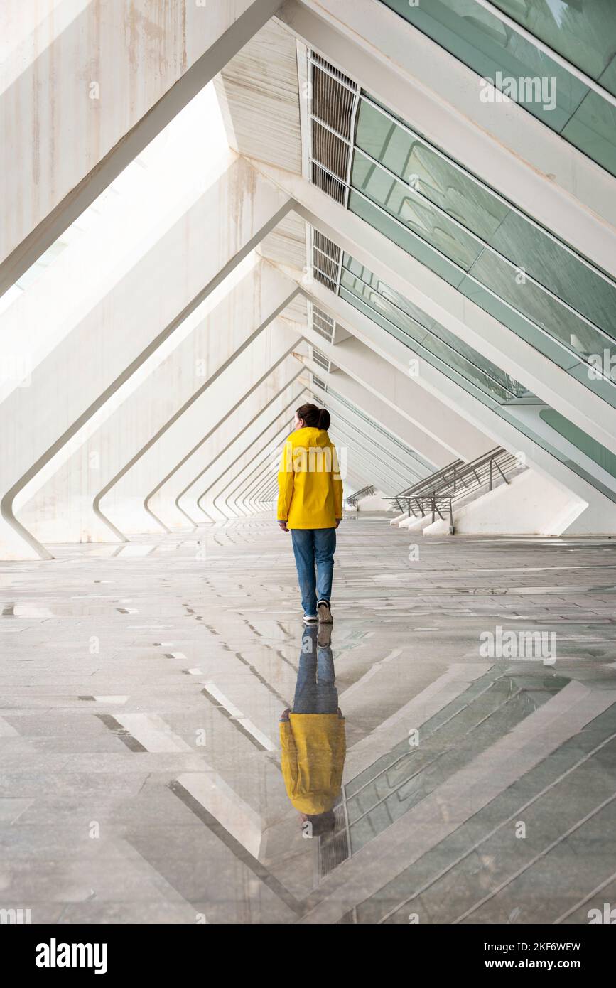 rear view of a person walking through modern architecture in the rain wearing a yellow coat. Stock Photo