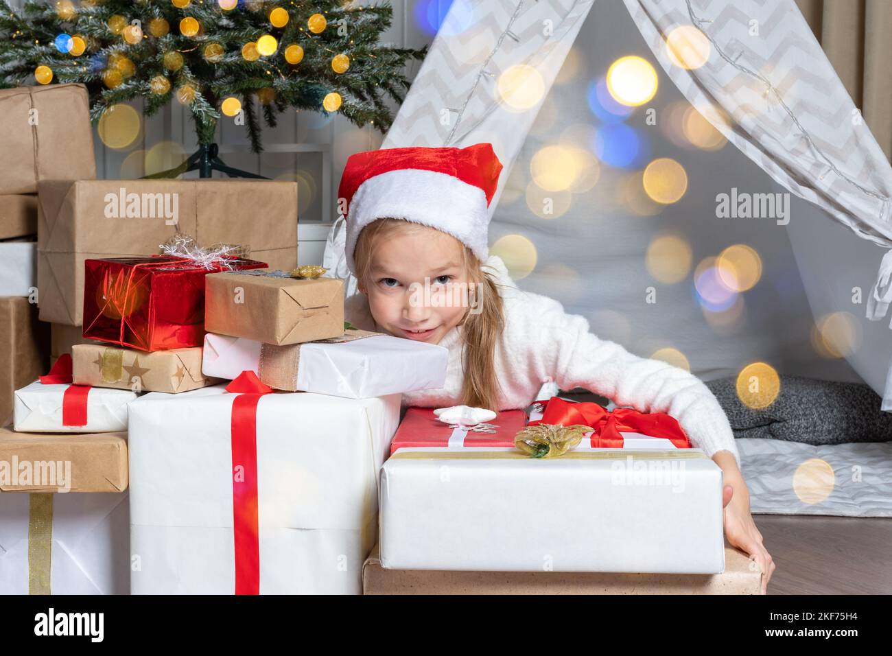 A pretty girl in a Santa hat holds her gifts for Christmas New Year lying in a children's room decorated with garlands, bokeh. Christmas gifts for chi Stock Photo
