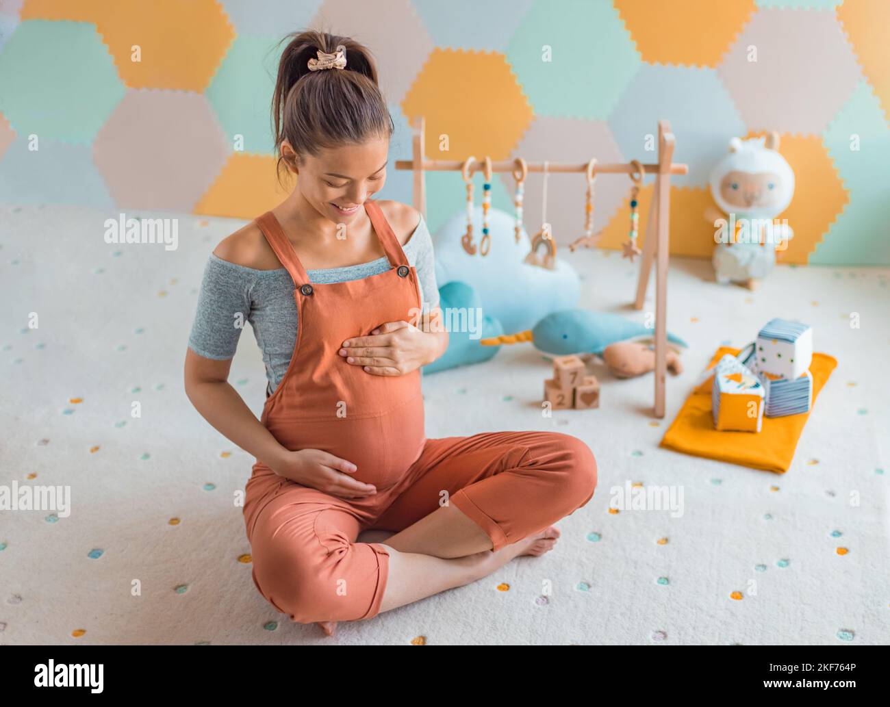 Pregnant woman smiling at belly in nursery playrooom with Baby activity gym play, toys and playmat. Pregnancy concept and home nusery planning and Stock Photo