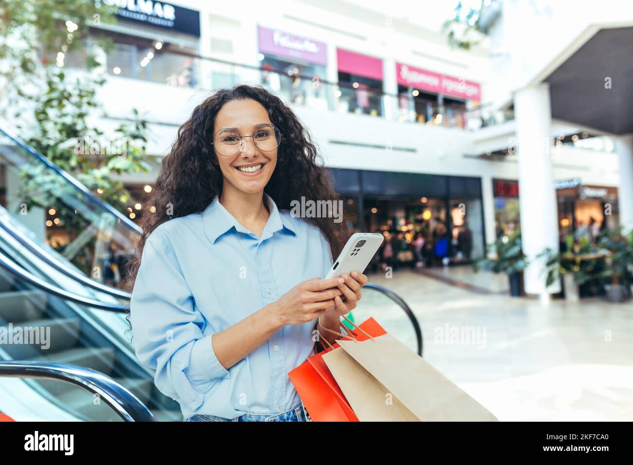 Business shopping. A young beautiful Latin American woman, a businesswoman stands in a shopping center near an escalator, holds a phone and paper bags with goods in her hands, makes purchases, smiles. Stock Photo