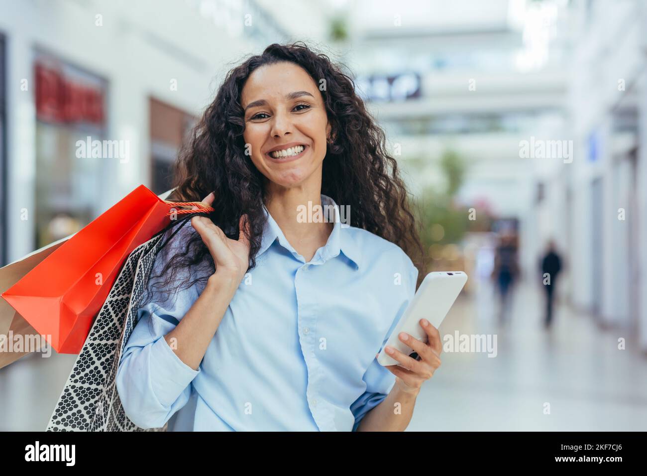 Portrait of happy hispanic woman shopper in store, looking at camera and smiling holding colorful shopping bags and gifts. Stock Photo
