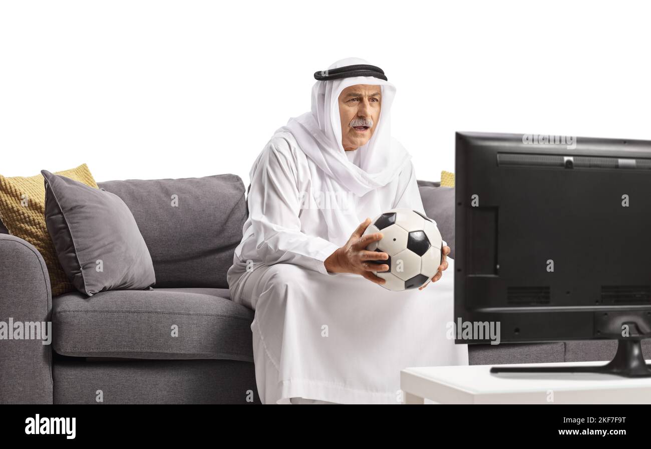 Mature arab man in a robe holding a football and sitting on a sofa and watching tv isolated on white background Stock Photo
