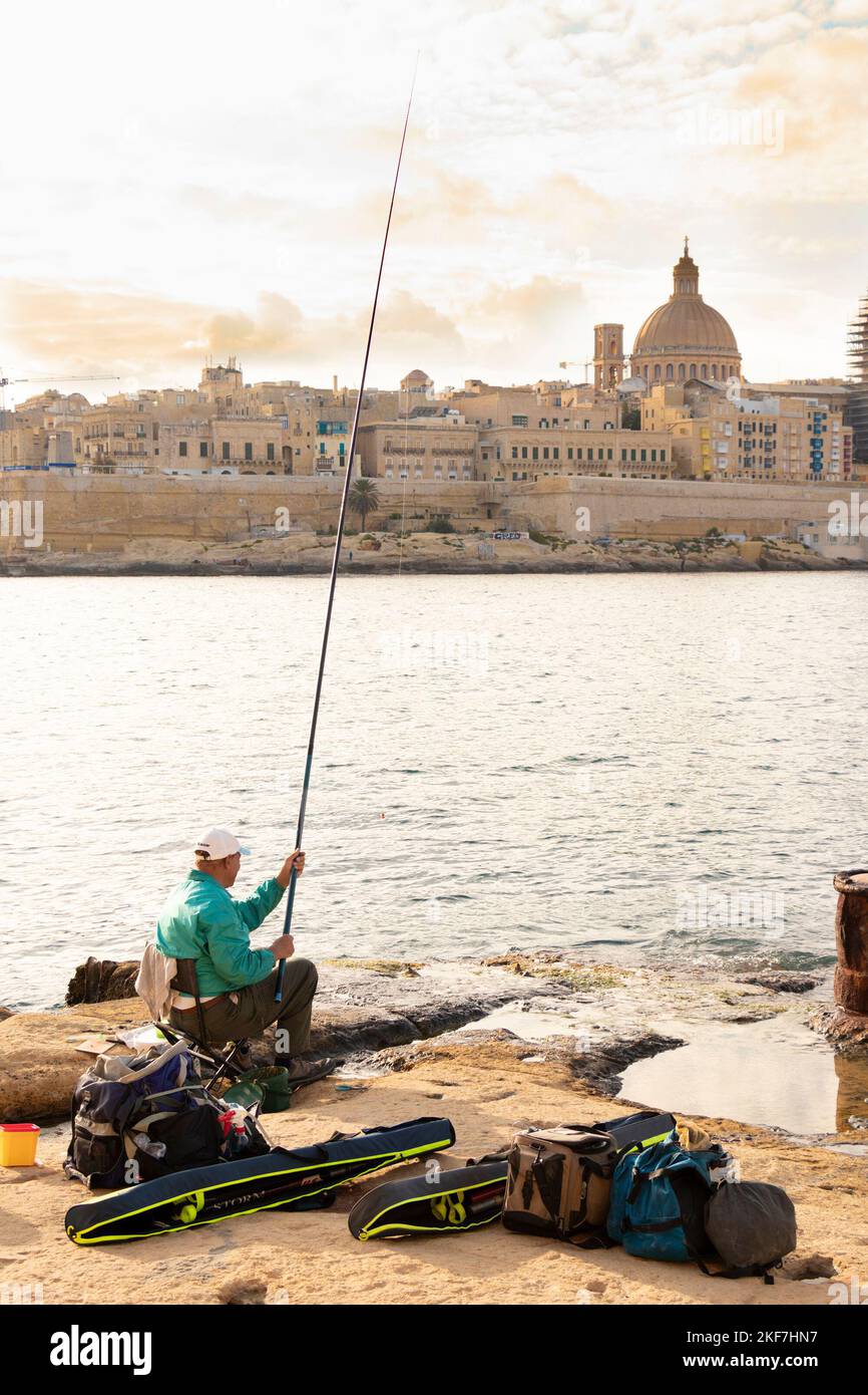 Sliema, Malta - November 12, 2022: Maltese fisherman sitting on a chair on the rocks by the sea while fishing with fishing stick on a sunny morning an Stock Photo