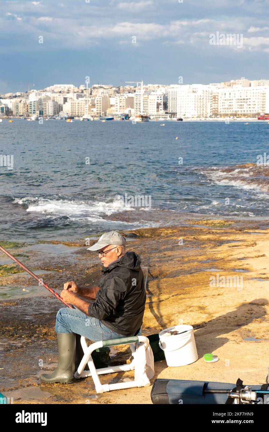 Sliema, Malta - November 12, 2022: Maltese fisherman sitting on a chair on the rocks by the sea while fishing with fishing stick on a sunny morning Stock Photo