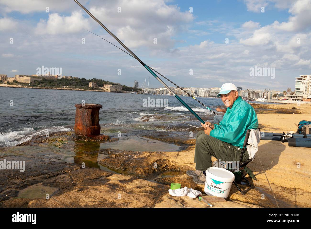 Sliema, Malta - November 12, 2022: Maltese fisherman sitting on a chair on the rocks by the sea while fishing with fishing stick on a sunny morning Stock Photo