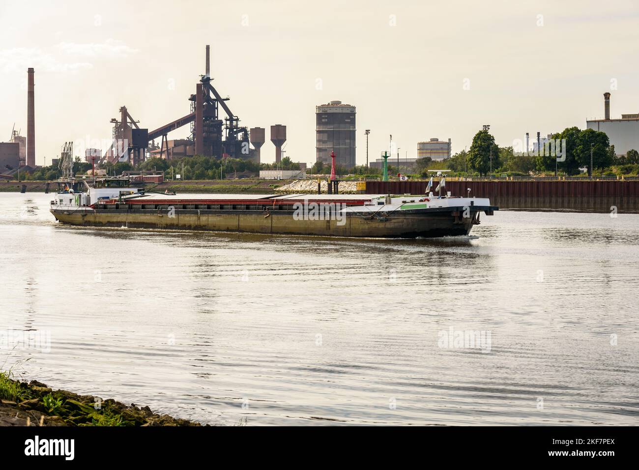 Cargo barge sailing up a river at sunset in summer. A riverside steel plant with a blast furnace and a gasometer is visible in background. Stock Photo