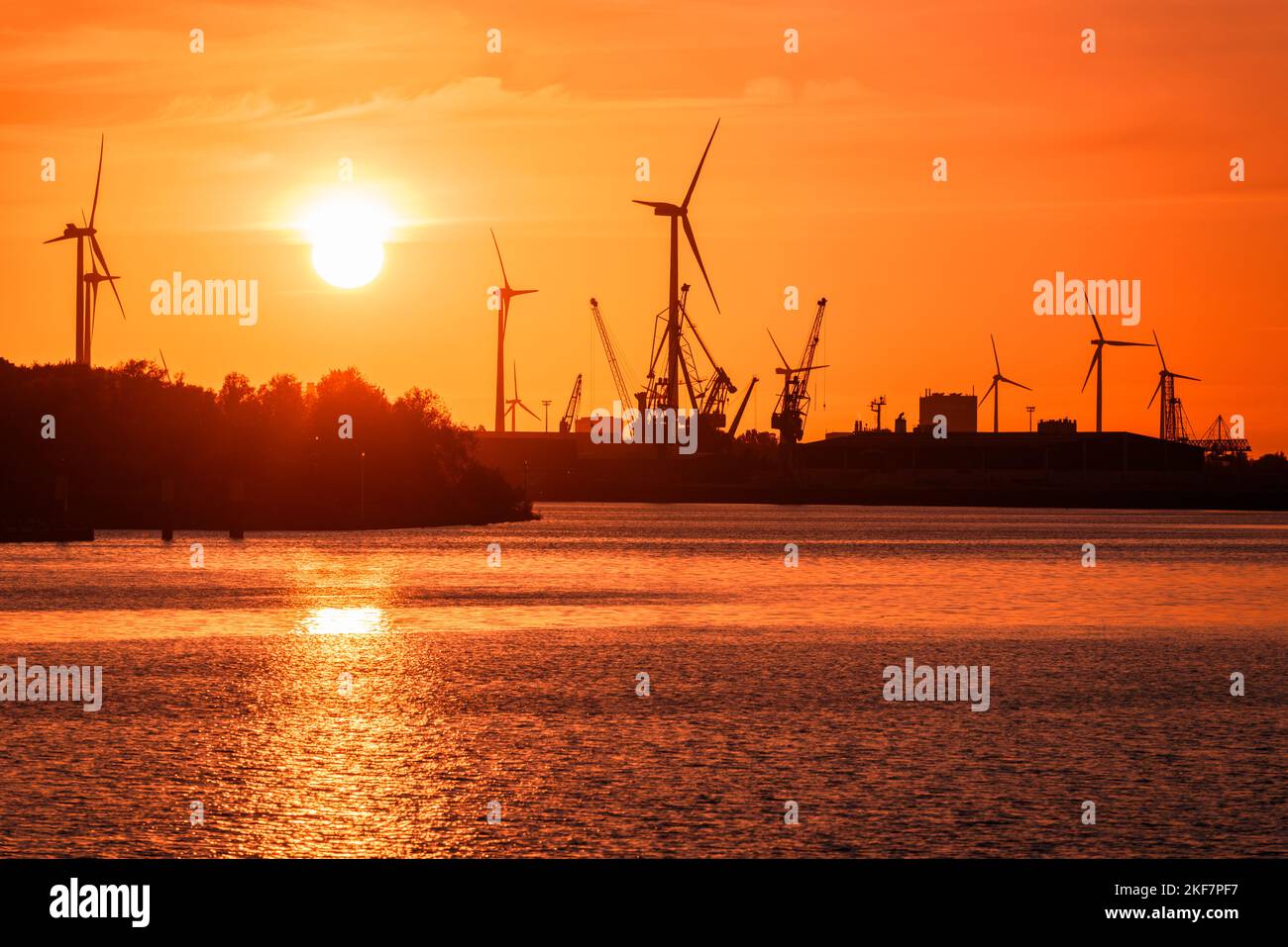 Industrial buildings along with wind turbines and dock cranes on a river harbour silhouetted against a fiery sky at sunset Stock Photo