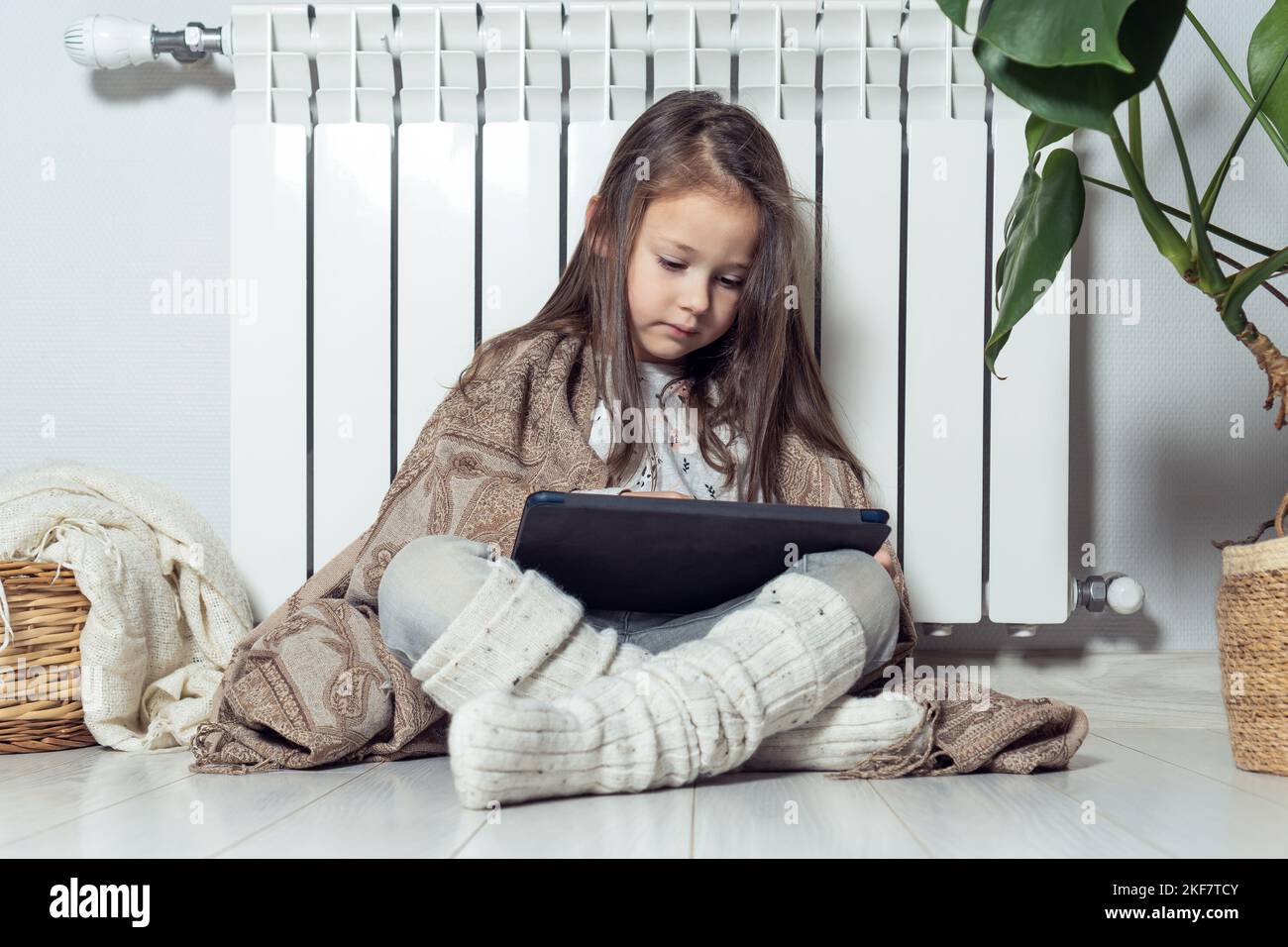 Small cute brunette long haired girl using electronic tablet, watch video, share messages, wrapped in blanket, sitting near warm heating radiator Stock Photo