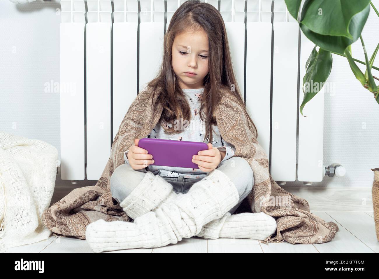 Small little brunette long haired girl using electronic tablet, surfing net and study online, wrapped in blanket, sitting near warm heating radiator Stock Photo