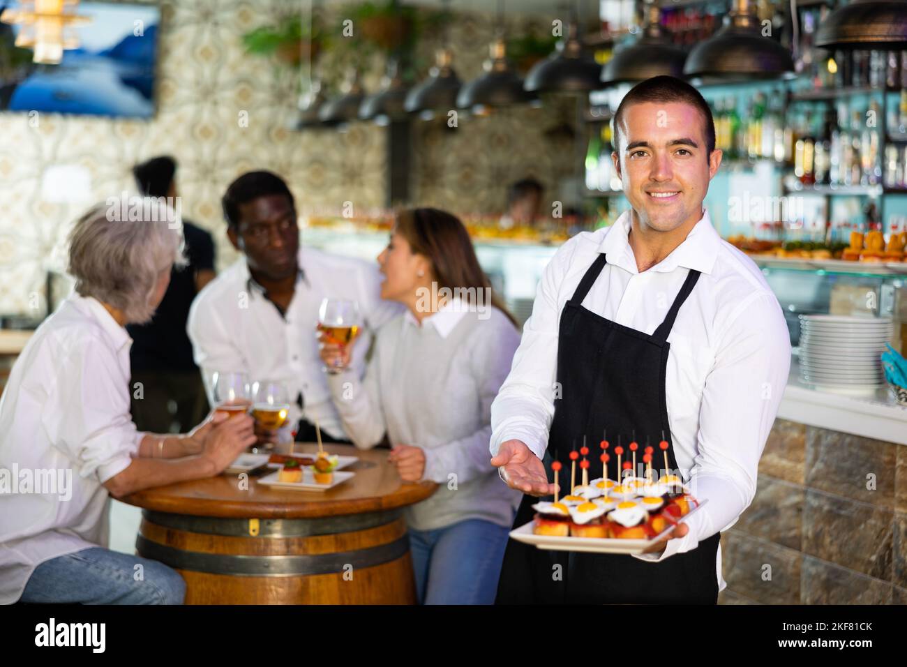 Smiling waiter posing and holding tray with order at bar with customers ...