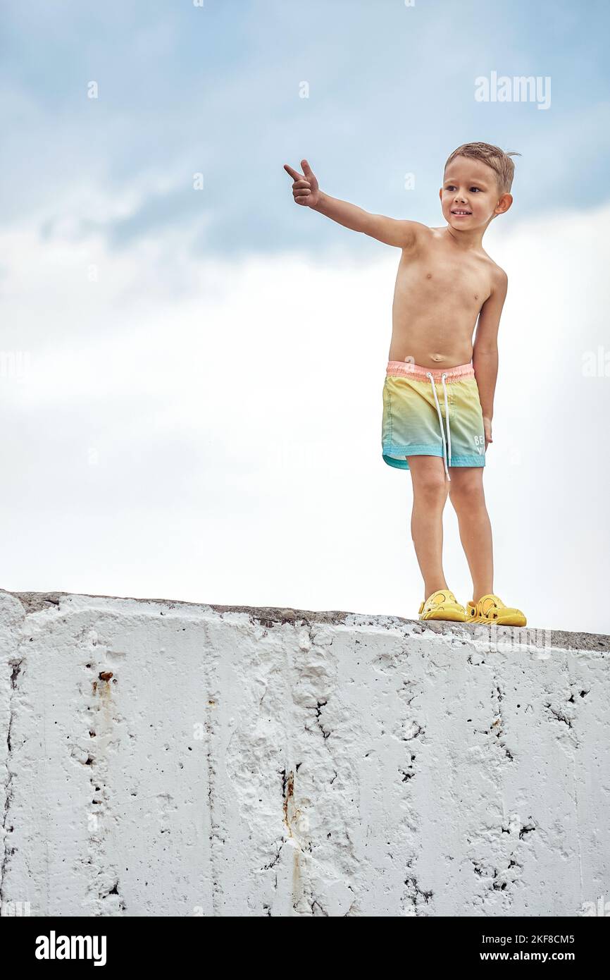Preschooler boy points finger to interesting thing standing on concrete pier of beach. Puzzled child explores sea nature on summer holidays Stock Photo