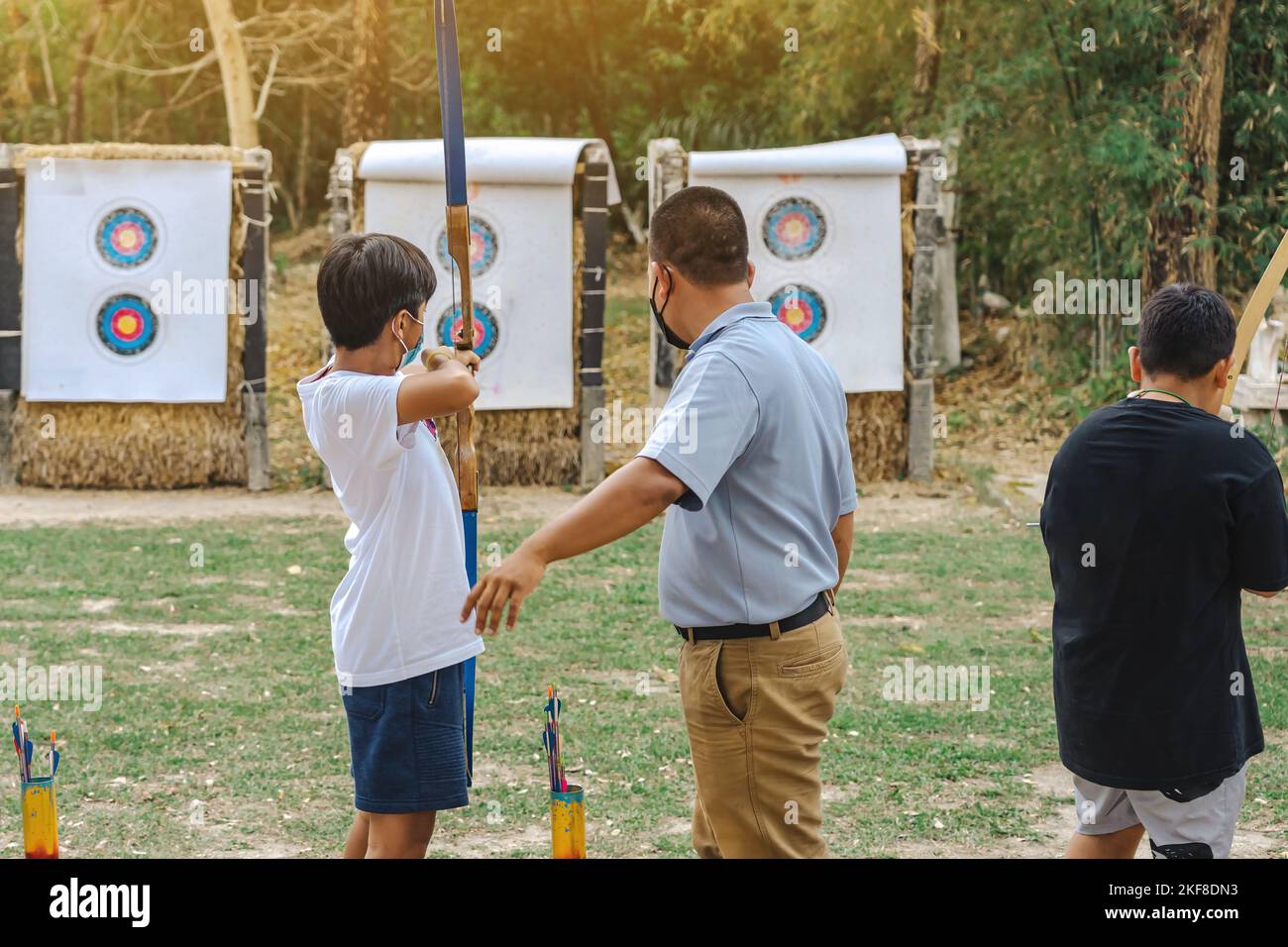 Back view of Asian boy wear face mask aims archery bow and arrow to colorful target in shooting range during training and competition. Exercise and co Stock Photo