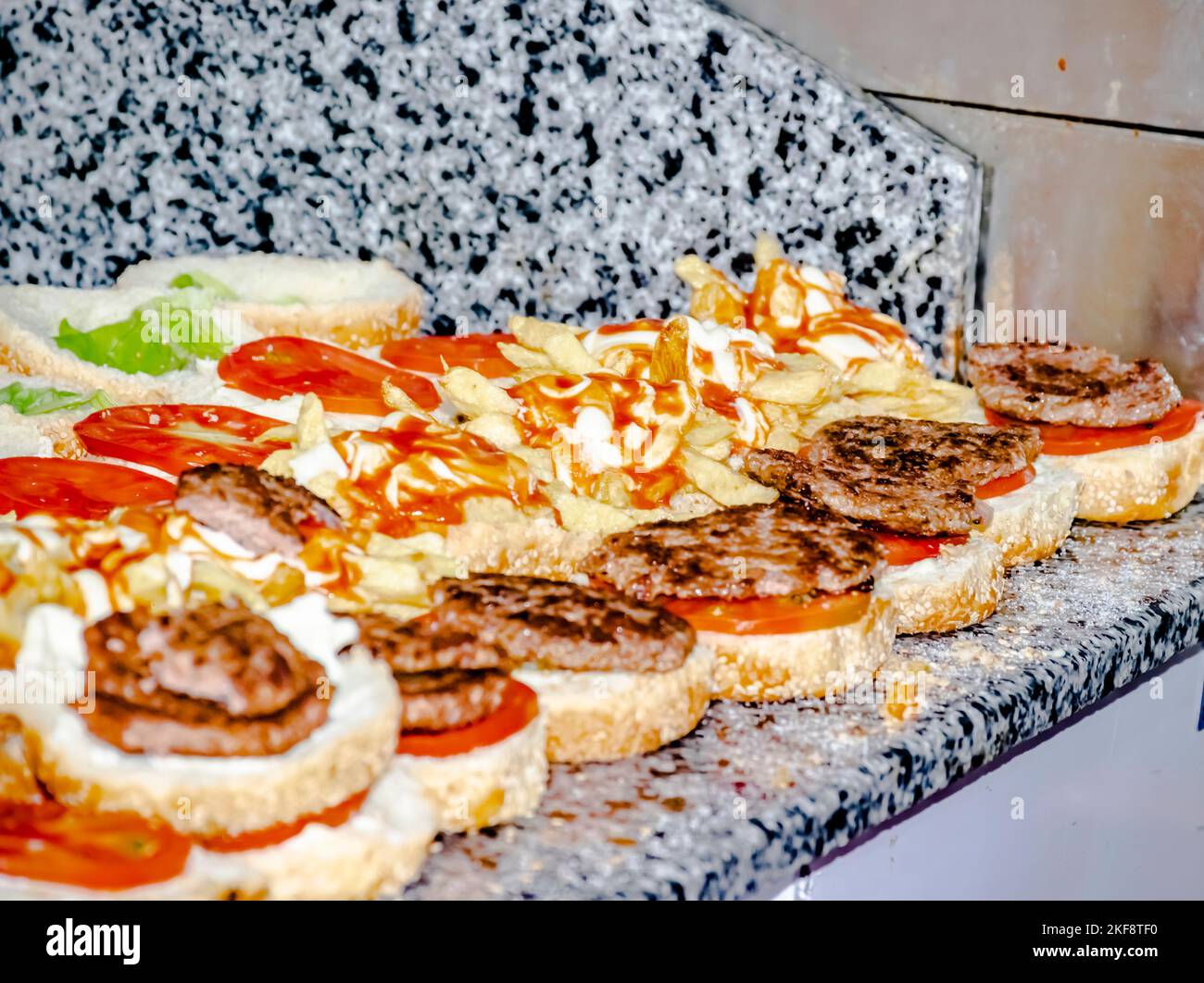 Burgers in a row on worktop, focus on opened hamburgers filled with french fries, lettuce and tomato, grilled chopped steaks, mayonnaise and ketchup. Stock Photo