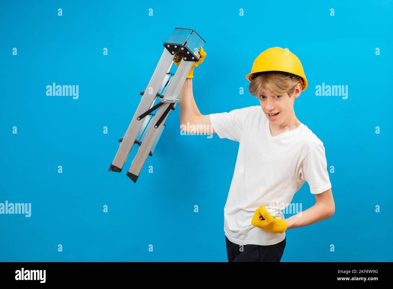 Young repairman teen in white t-shirt and gloves with yellow helmet in head, holding ladder in hands standing in studio on blue background. Stock Photo