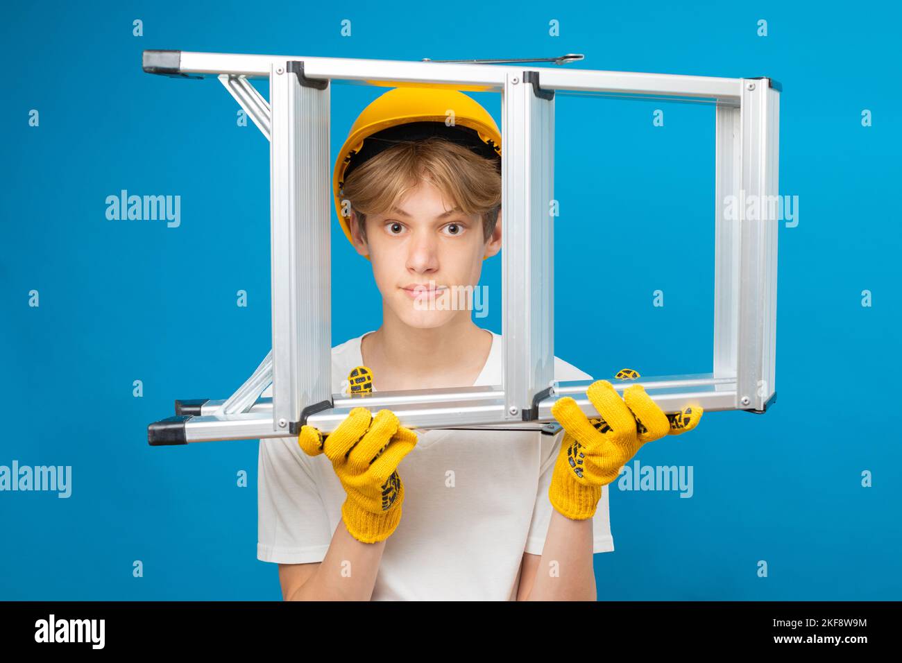 Young repairman teen in white t-shirt and gloves with yellow helmet in head, holding ladder in hands standing in studio on blue background. Stock Photo