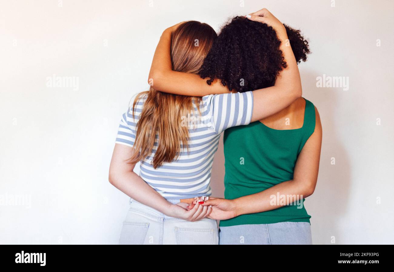Strong female friendship. Rear view two teen girls best friends holding hands behind back and hugging while standing in front of beige wall Stock Photo