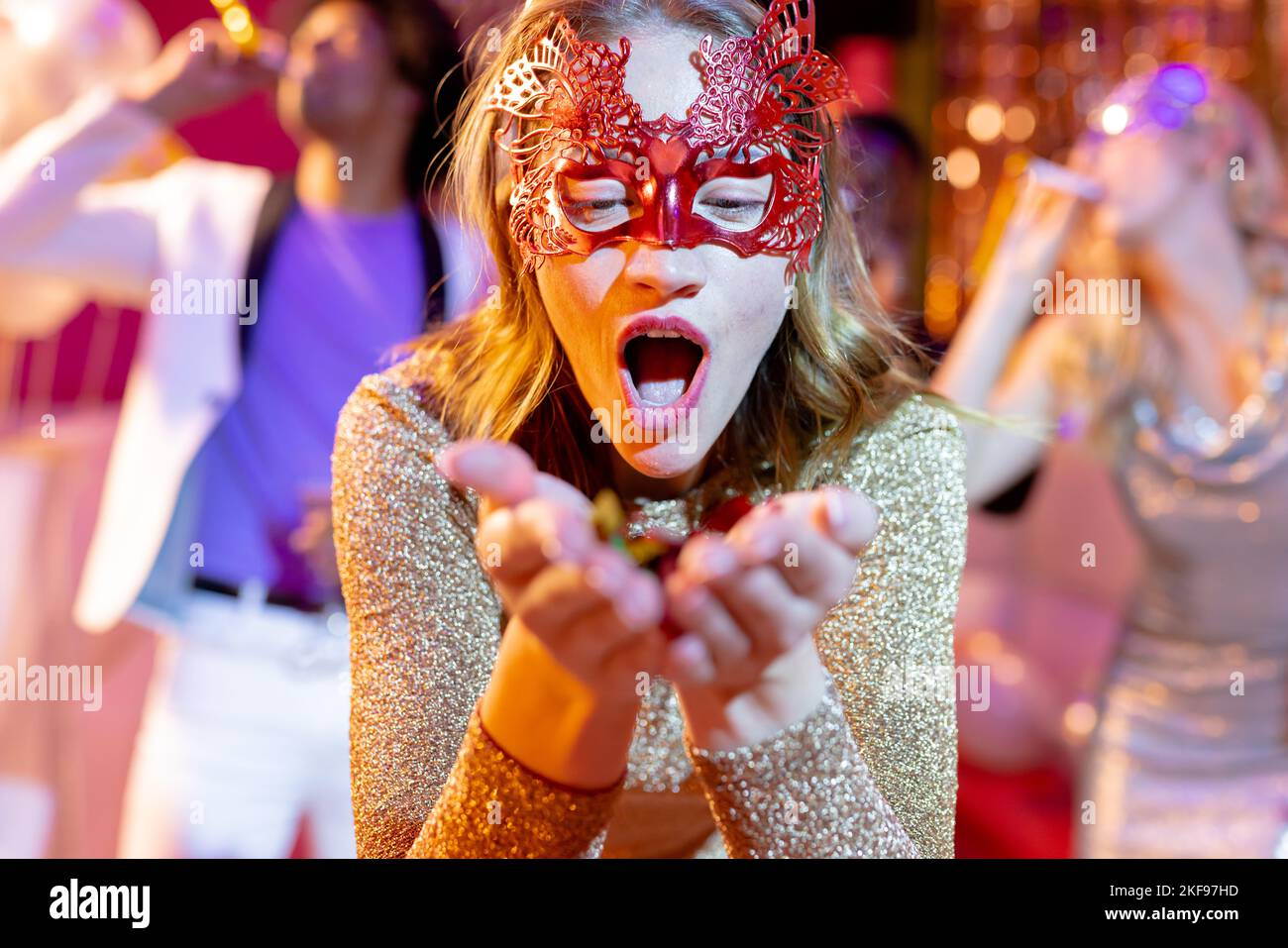 Happy caucasian woman in mask blowing glitter on the dancefloor at a party in a nightclub. Fun, going out, celebrating and party concept. Stock Photo