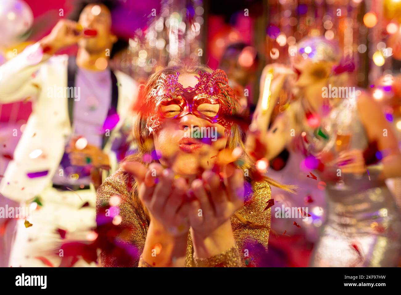 Happy caucasian woman in mask blowing glitter to camera at a party in a nightclub. Fun, going out, celebrating and party concept. Stock Photo